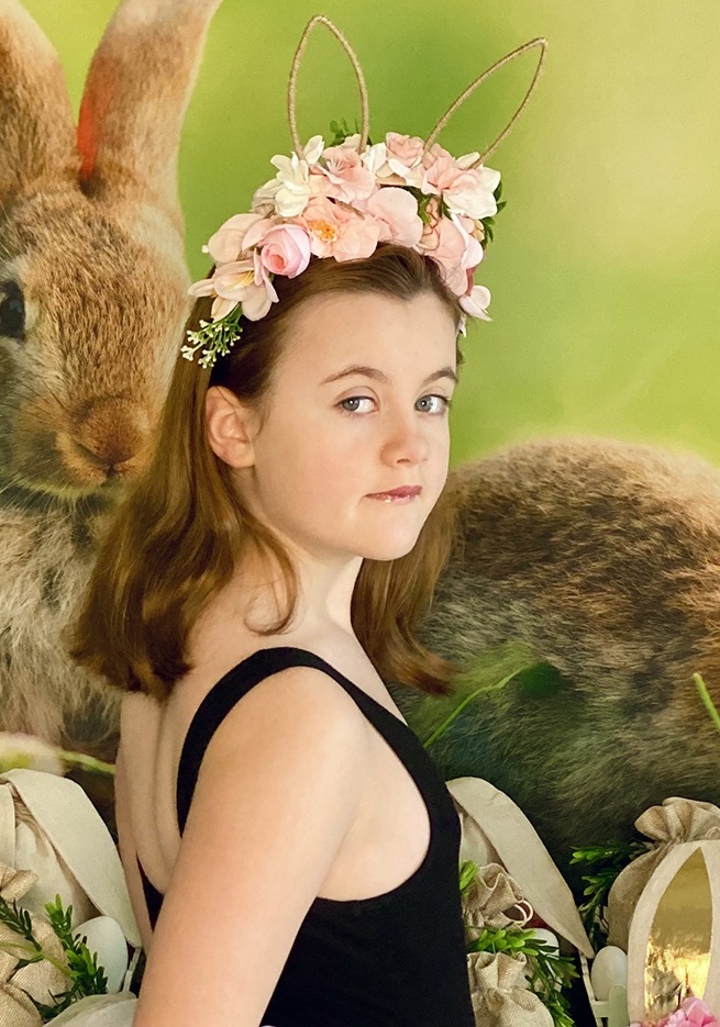 Close-up of young girl looking back over her shoulder with a gentle, thoughtful expression in front of Easter fairy garden party setup.