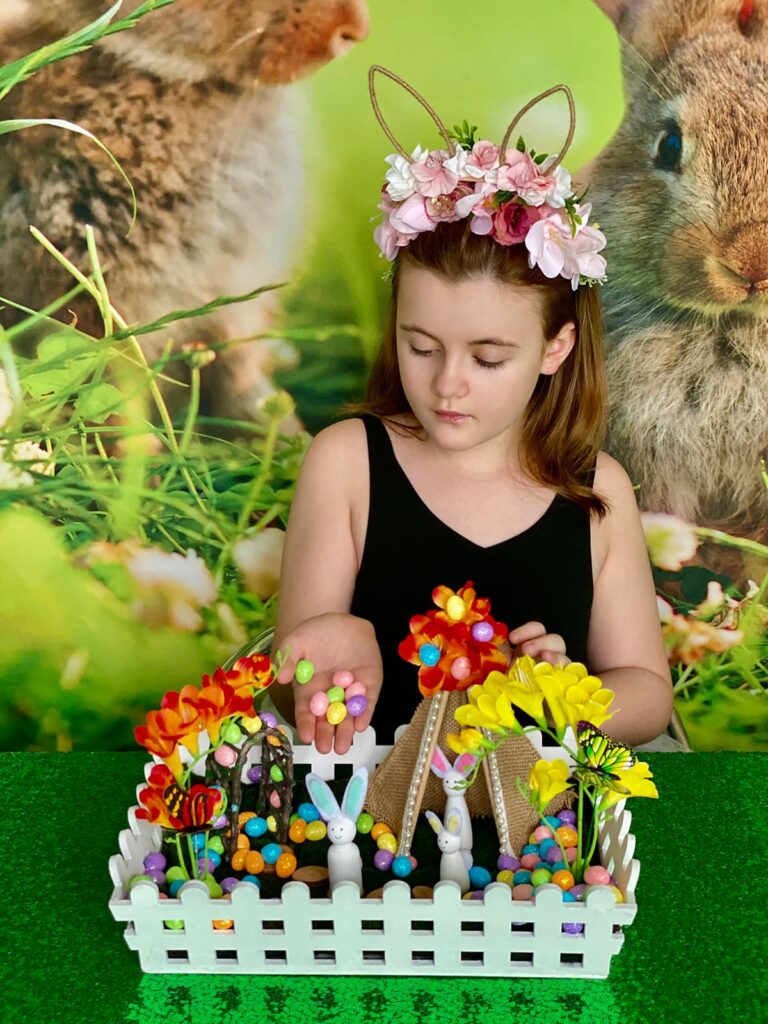 Child carefully scattering pastel foam eggs into the Easter fairy garden crate, eggs rolling off her hand as she looks down thoughtfully.