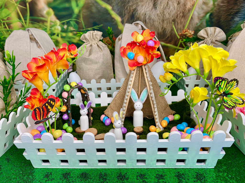 Detailed image of a completed Easter fairy garden inside a white picket crate, featuring miniature bunnies, faux moss, tiny flowers, teepee and stepping stones arranged in a magical spring scene.