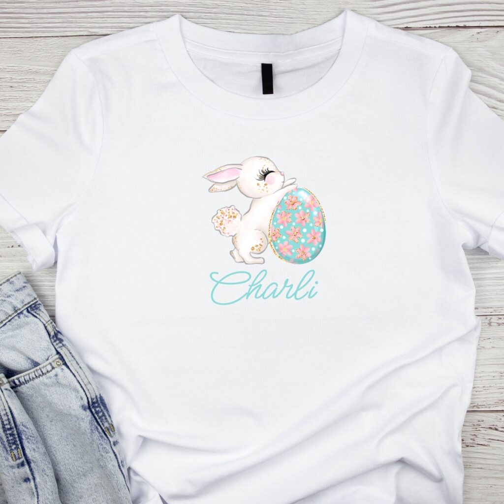 Custom Easter t-shirt featuring a bunny, pastel Easter egg, and a child’s name in personalized lettering, available from Inspired Party Merch.