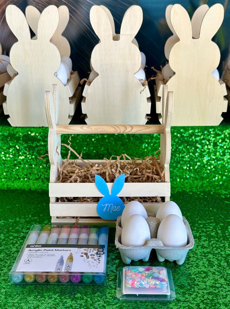 Bunny-shaped wooden crate filled with ceramic Easter eggs, metallic paint markers, and adhesive gems as a DIY Easter egg decorating party favour for kids.