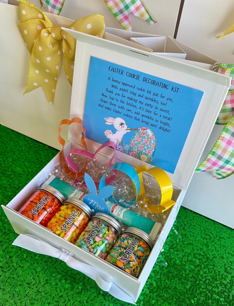 Easter cookie decorating kit party favor box filled with colorful Easter-themed sprinkles, bunny and egg-shaped cookie cutters, pastel icing pens, and a cookie recipe card for kids to bake at home.