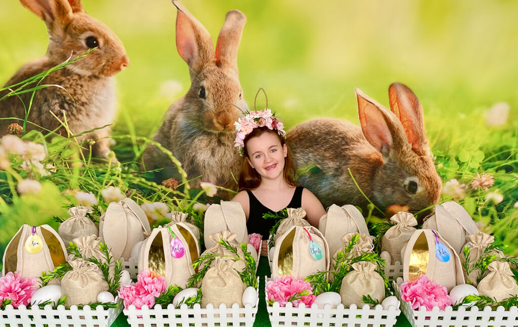 Wide view of Easter fairy garden party activity table with a child smiling behind it, featuring decorated crates, bunny backdrop, and spring-inspired setup.