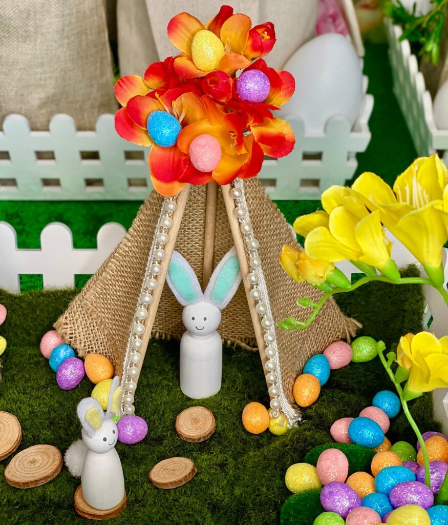 Easter Fairy teepee with wooden bunnies inside and on wooden stepping stones, surrounded by pastel eggs and a fairy crate filled with vibrant orange and yellow flowers.