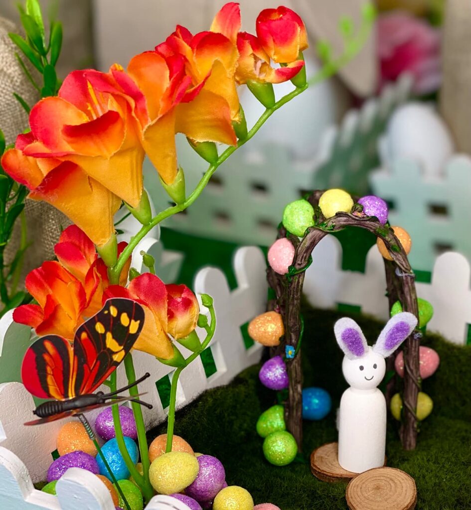 Close-up of a wooden Easter bunny standing on a wooden stepping stone beneath a fairy arch, surrounded by faux orange flowers and pastel Easter eggs in a fairy garden crate.