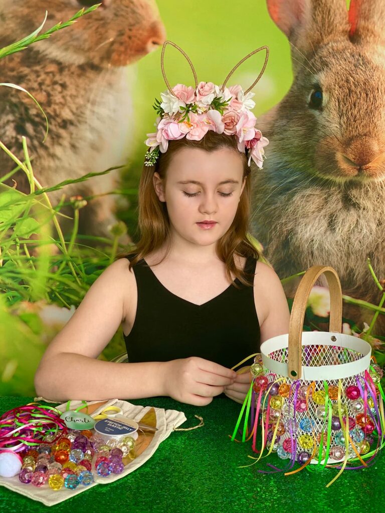 Girl peacefully tying a piece of ribbon to her nearly finished Easter basket. Supplies are neatly arranged beside her on the bunny ear bag.