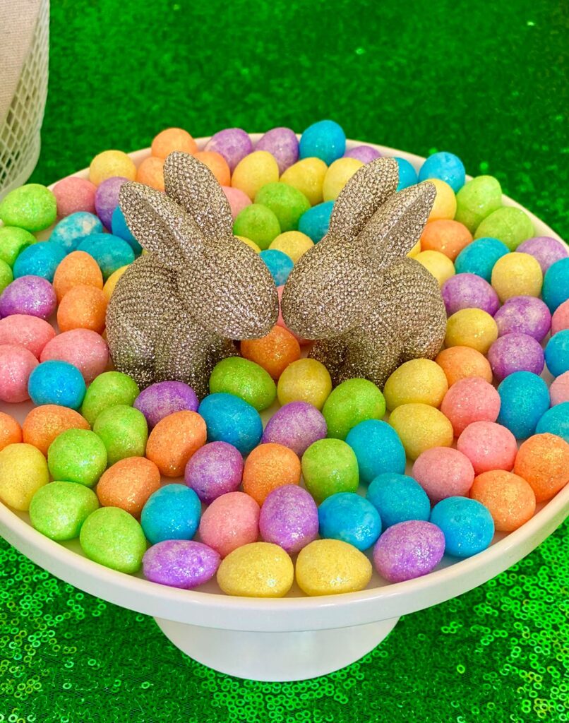 Top-down view of a white cake stand with gold bunny decorations surrounded by pastel eggs, used as a centerpiece for an Easter activity table.