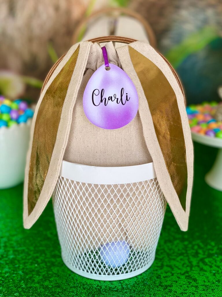 Close-up of a white mesh Easter basket with a bunny ear bag filled with basket decorating supplies, a fluffy cottontail, and a purple egg-shaped name tag labelled Charli.