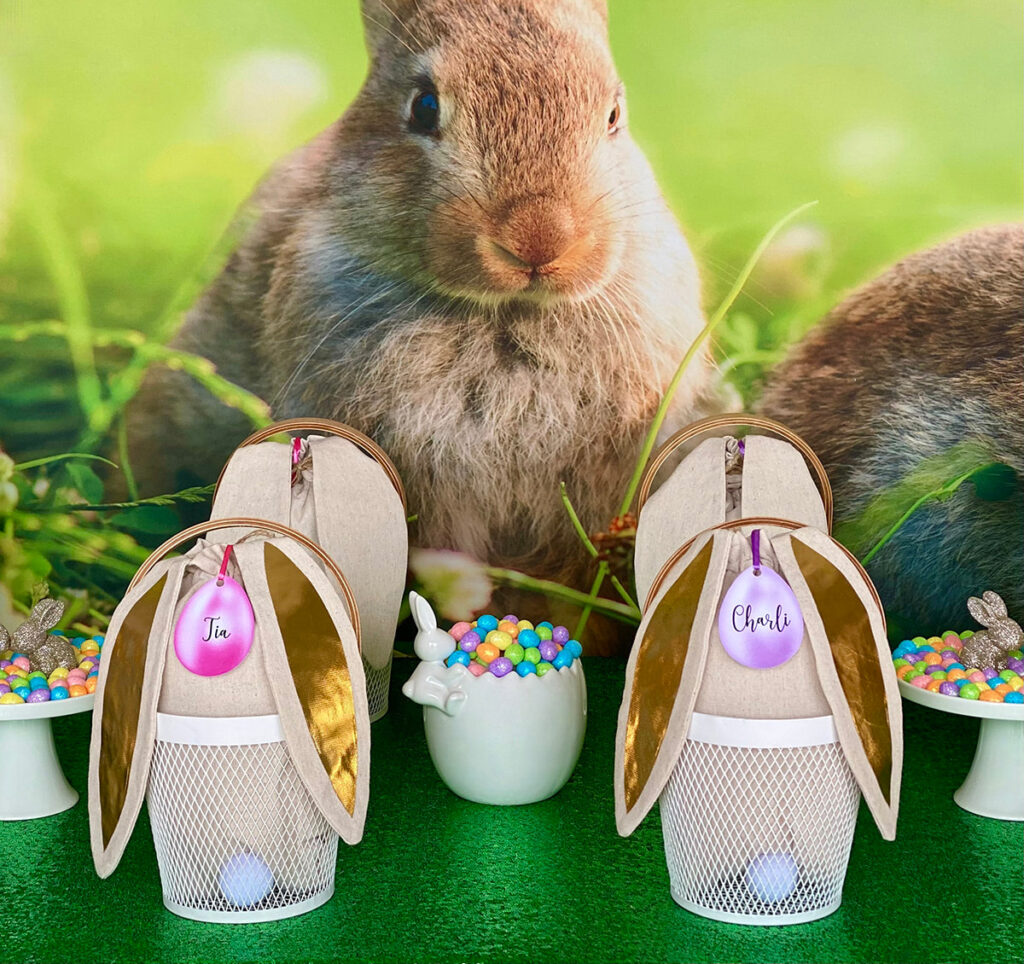 Close-up of two white mesh Easter baskets with gold bunny ear bags filled with decorating supplies, each labelled with a personalised pastel name tag, set on a green table with candy dishes and a bunny backdrop.
