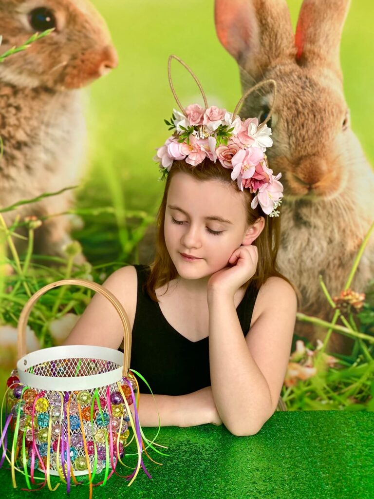 Girl smiling sweetly at the decorated Easter basket she has made, resting her elbow on the table with her hand on her face, looking at the basket innocently.