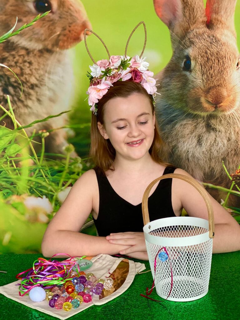 Smiling girl with arms crossed in front of her, admiring a bead and ribbon she attached to a white wire Easter basket. Decorating supplies are neatly displayed on the table beside her.