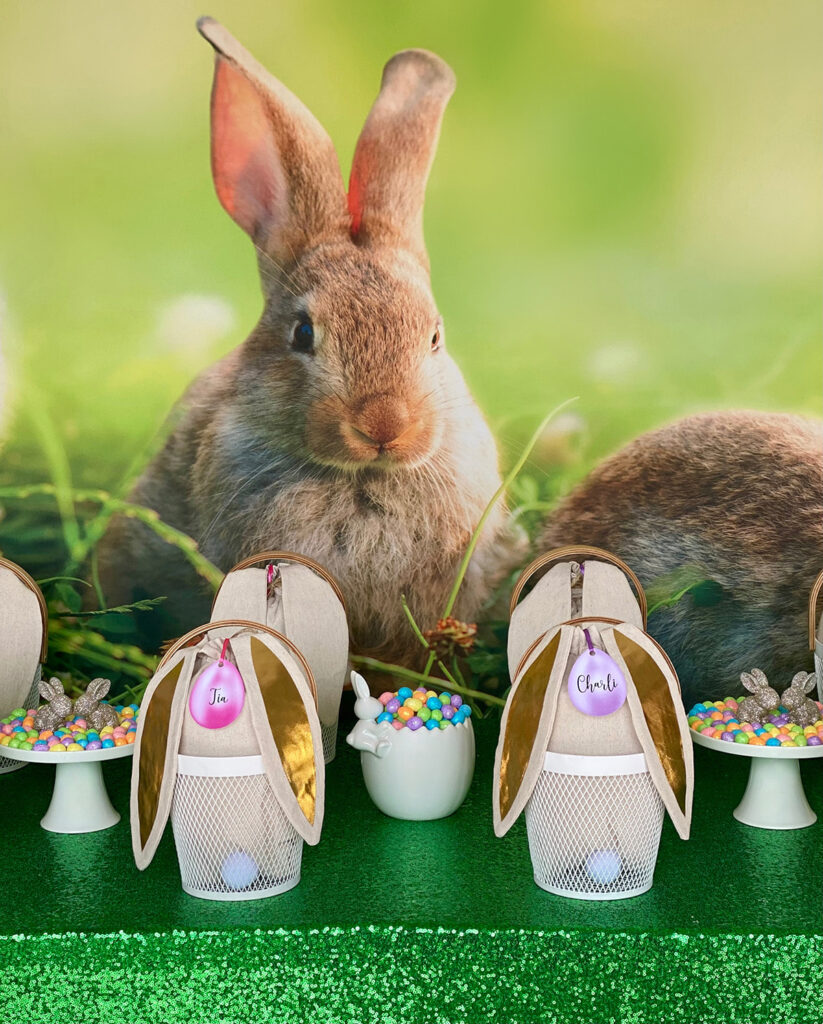 Kids’ Easter party table setup featuring personalised white mesh baskets with bunny ear bags filled with decorating supplies, pastel egg candy dishes, a green sequin tablecloth, and a spring bunny backdrop.