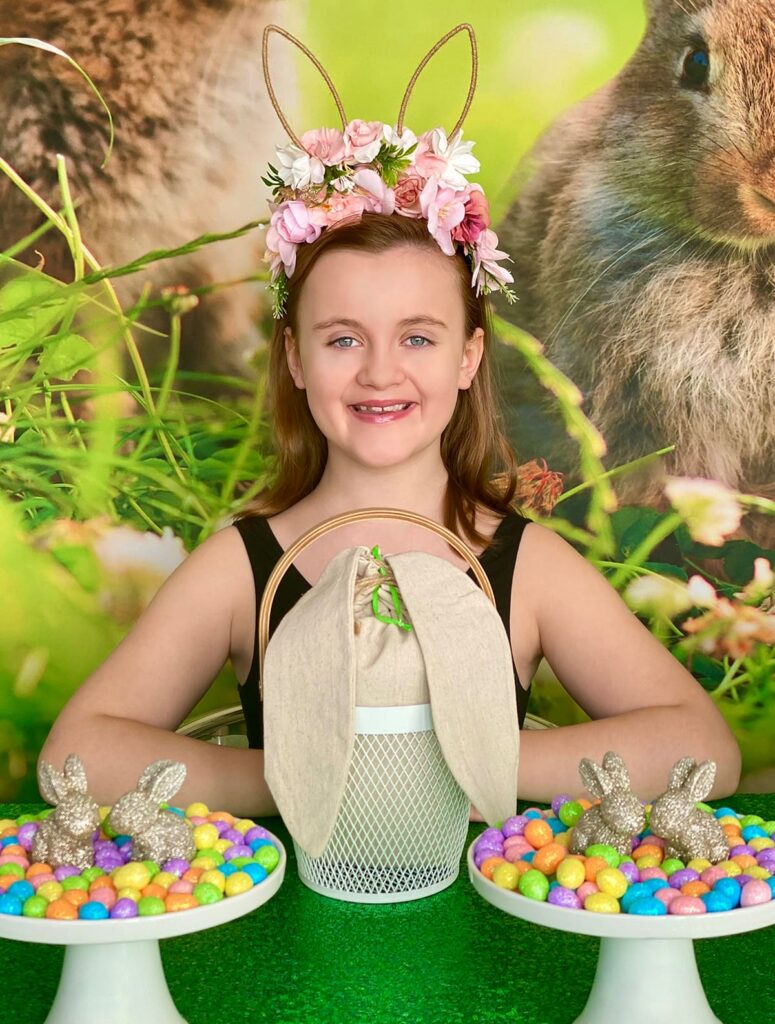 Smiling girl sitting at an Easter basket decorating table with her basket and bunny ear bag in front of her. White cake stands with gold bunnies and pastel eggs sit on a green sequin tablecloth, with a bunny backdrop behind her.