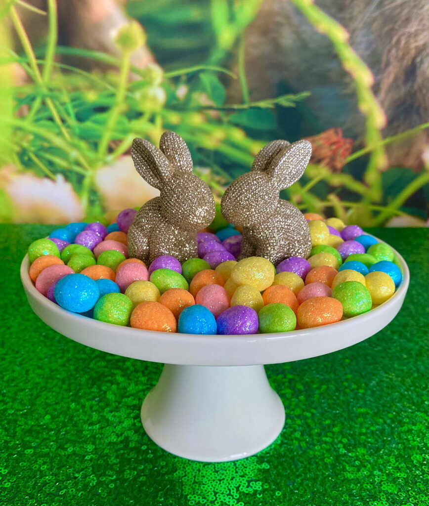 Close-up of a white cake stand on a green sequin tablecloth, featuring two gold sparkly bunny decorations surrounded by pastel eggs, with a spring bunny backdrop behind.
