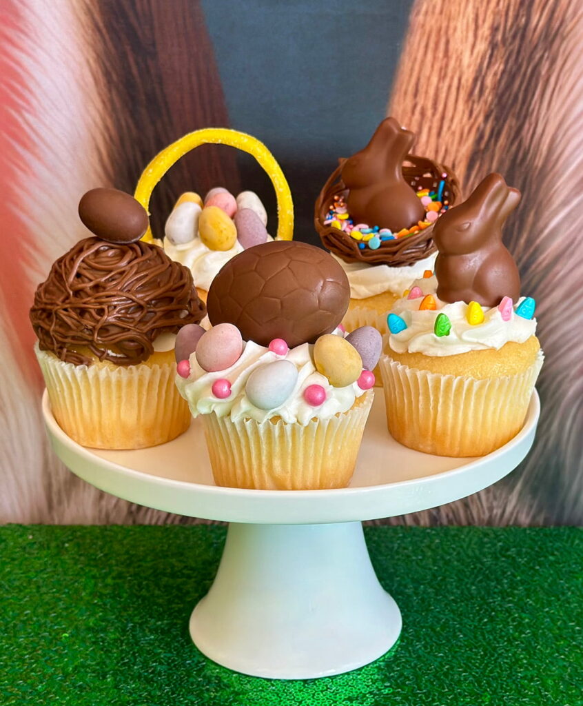 Assortment of Easter cupcakes displayed on a white cake stand, with a bunny ear backdrop behind and a green sequin tablecloth underneath.