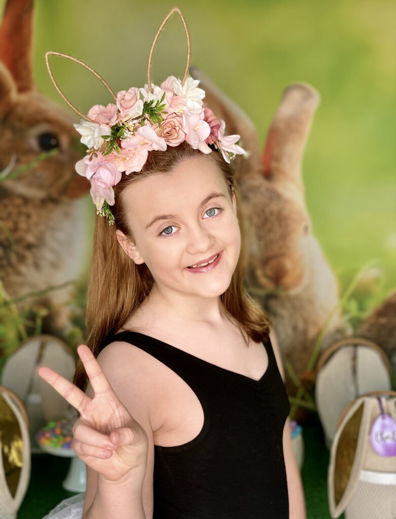 Happy girl smiling from ear to ear, wearing a floral bunny ear headband and making a peace sign with her fingers for the camera.