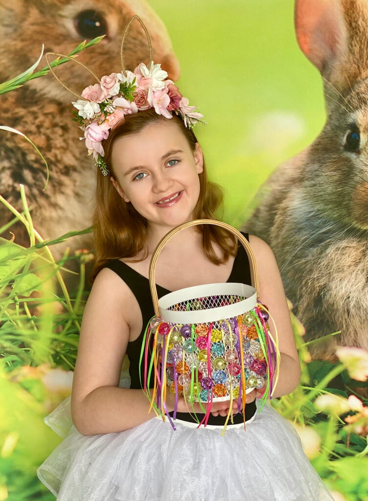 Happy girl holding her completed Easter basket while standing in front of a bunny-themed spring backdrop.