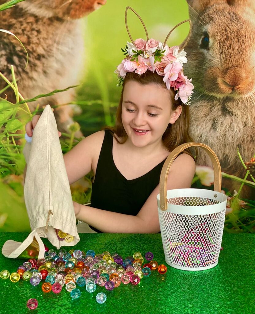 Smiling girl pouring colorful crystal beads from a bunny ear bag onto a green sequin tablecloth at an Easter basket decorating party.