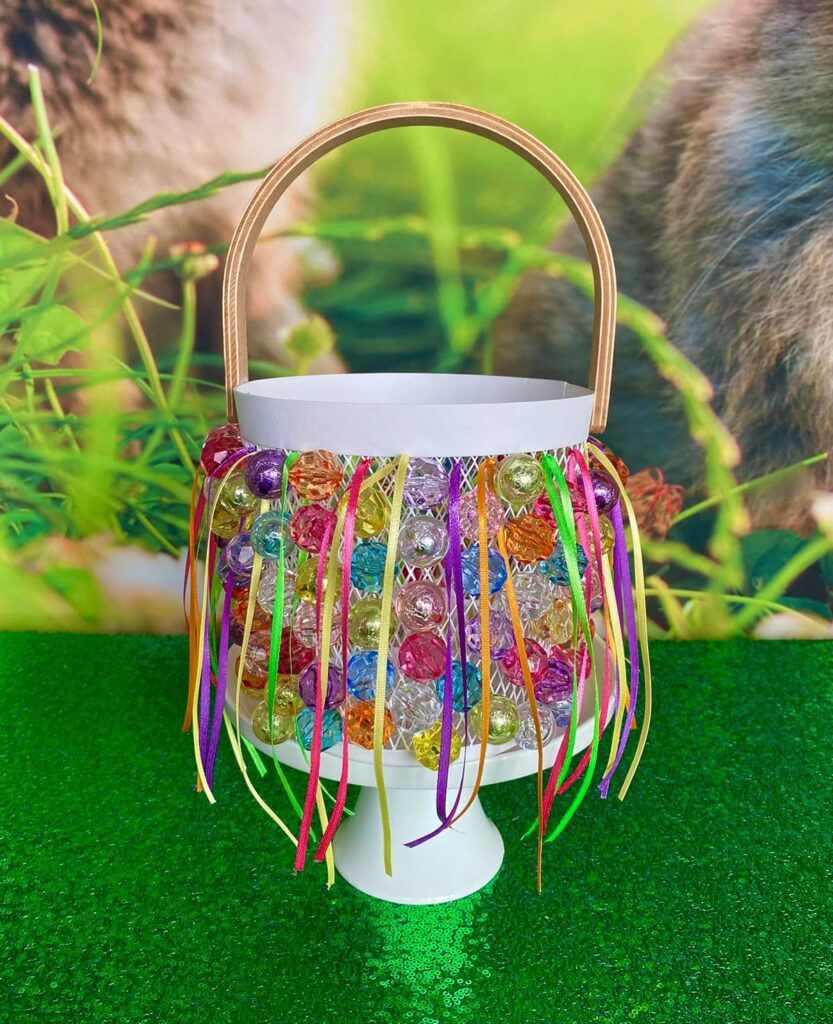Completed Easter basket displayed on a white cake stand atop a green sequin tablecloth, with a hint of the spring bunny backdrop in the background.