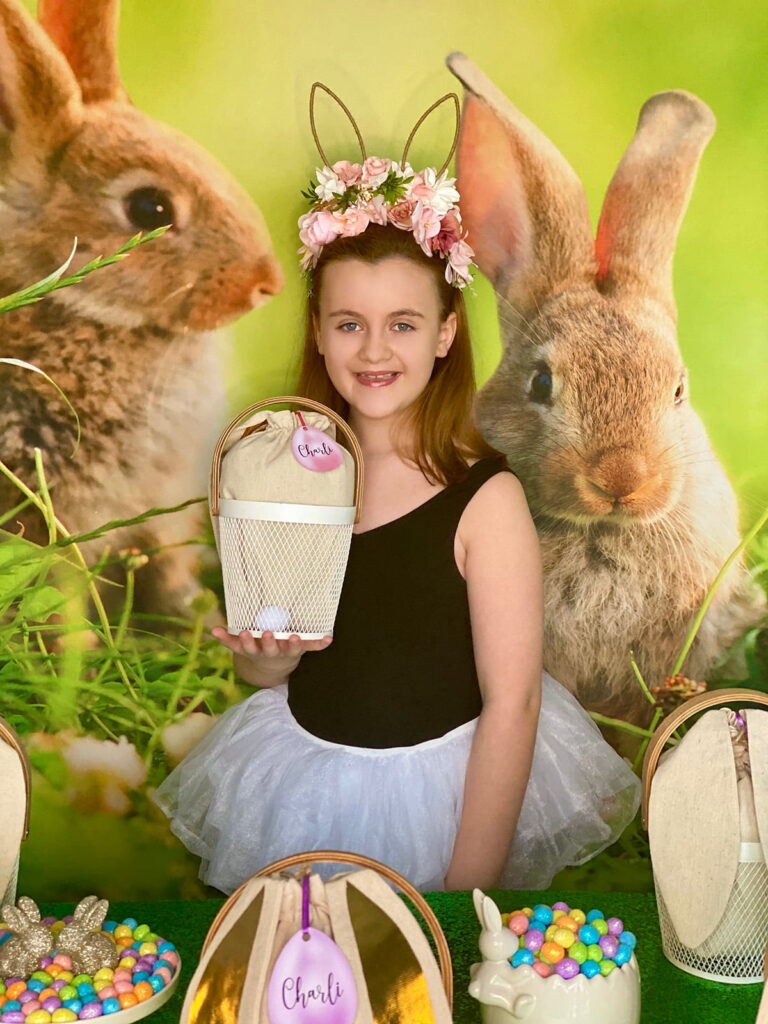Young girl smiling while holding a white mesh Easter basket with a bunny ear bag full of decorating supplies and a personalized Charli name tag, standing behind the party activity table with a spring bunny backdrop.
