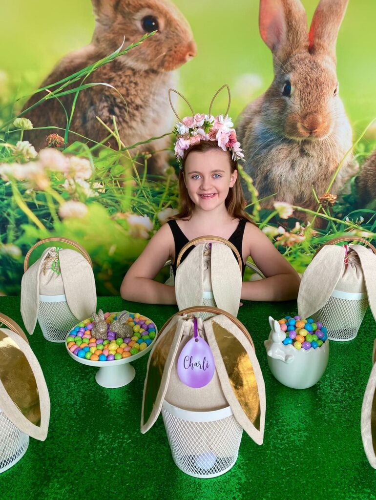 Young girl seated behind the Easter basket decorating activity table with her arms crossed on the table, smiling and ready to begin the craft, with bunny baskets and pastel decor in view.