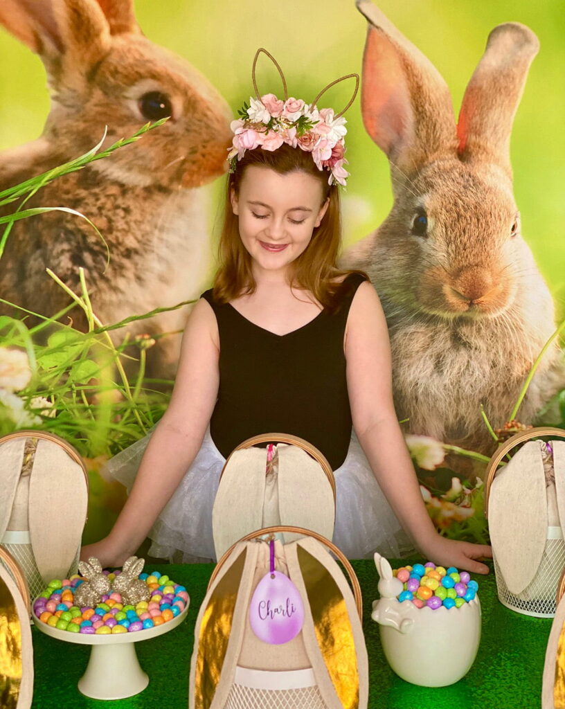Smiling girl standing behind an Easter basket decorating activity table, looking down at the setup with personalised activity baskets and pastel eggs with a spring bunny backdrop.
