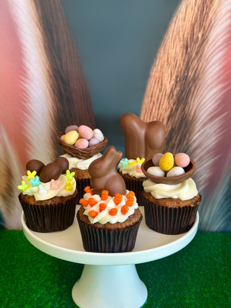 White cake stand holding five decorated Easter cupcakes topped with chocolate bunnies, Easter sprinkles, and chocolate nests filled with mini eggs, set on a green sequin tablecloth.