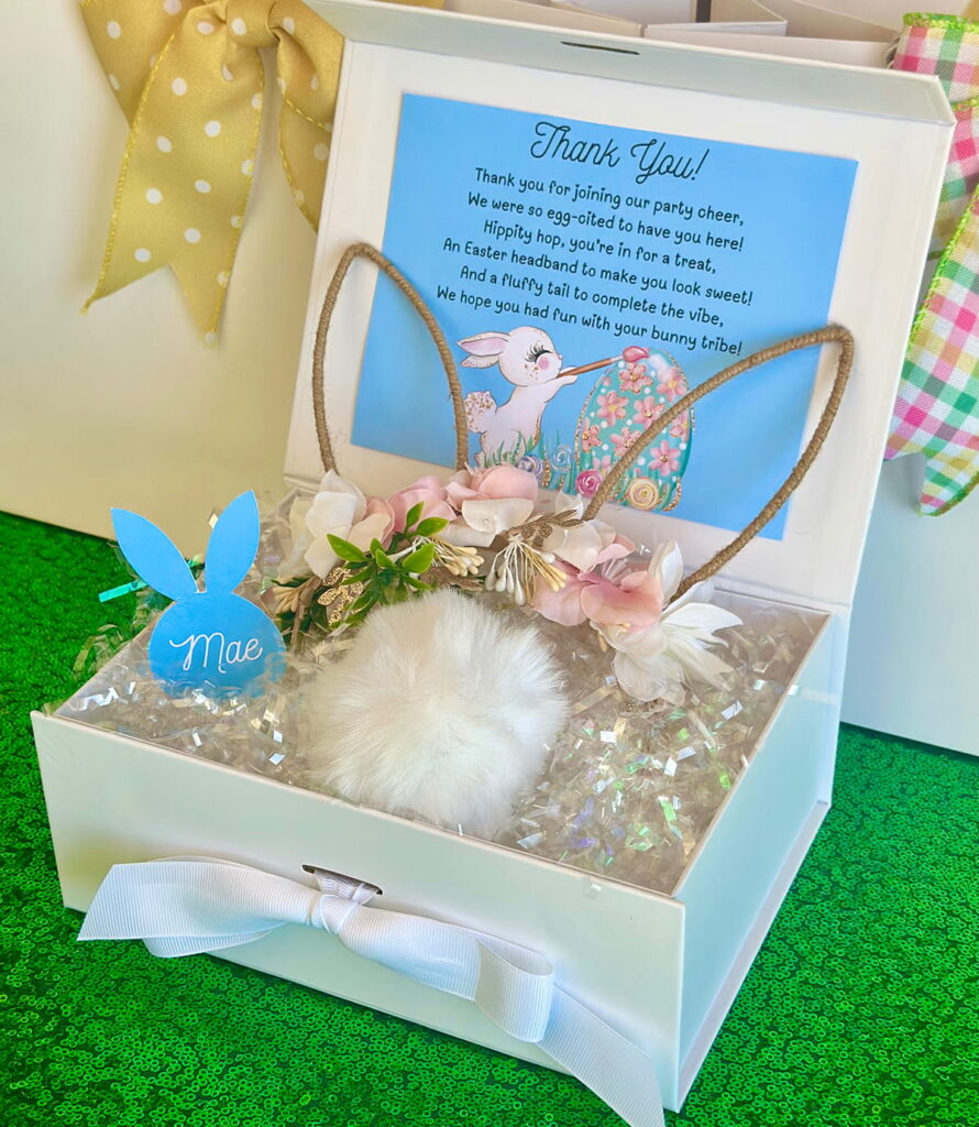 Top-down front angle of bunny accessories party favour box with floral bunny ear headband and white cottontail on iridescent shred inside a white magnetic gift box, featuring a blue bunny-shaped name tag and matching thank you card on the lid interior,