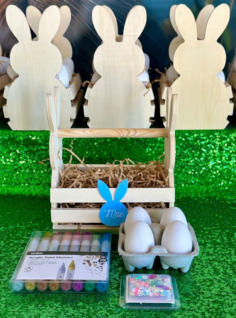 Top-down view of DIY Easter egg decorating kit with ceramic eggs, metallic paint pens, and pastel gems on green sequin tablecloth, in front of a bunny-shaped wooden crate with blue name tag and filled with brown shredded paper. Other crates are visible in the background.