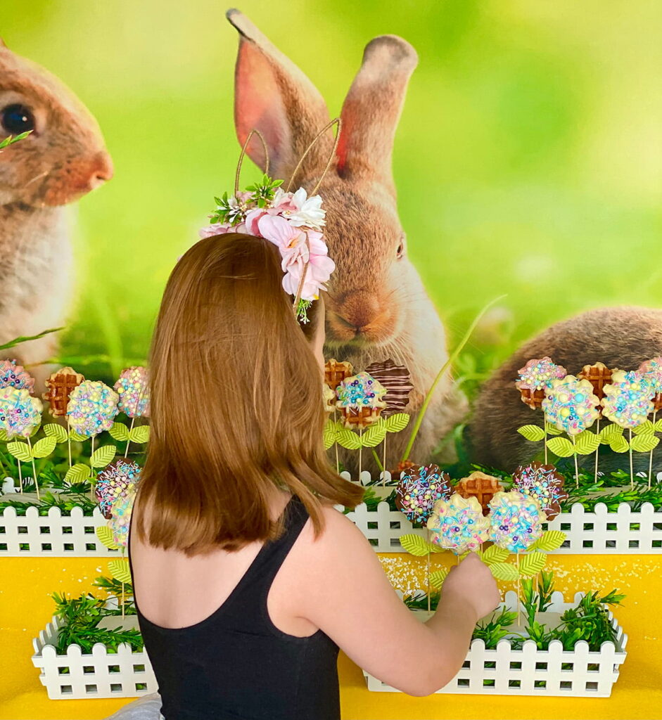 Young girl reaching for a waffle pop at a spring-themed Easter party table, with floral waffle pops, a bunny backdrop, and a yellow sequin tablecloth visible behind her.