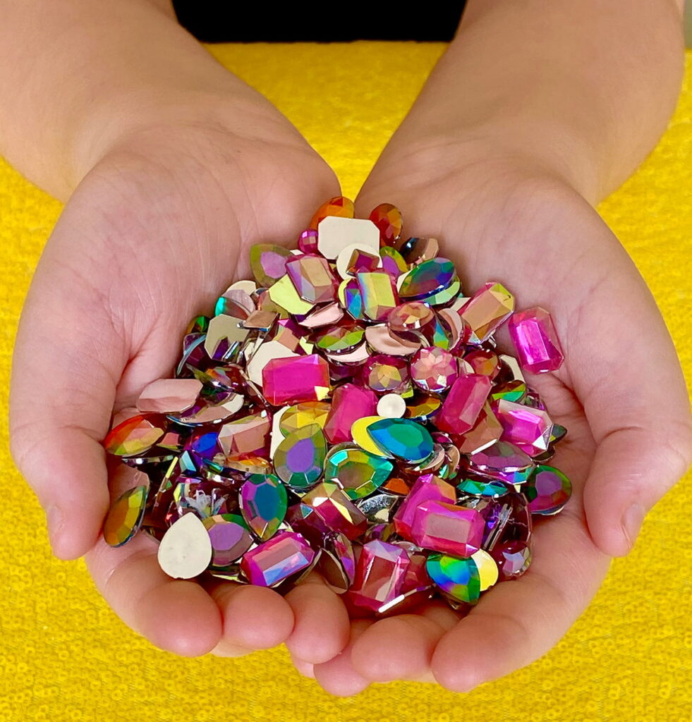 Colorful acrylic gems resting in the palms of hands over a yellow sequin tablecloth.