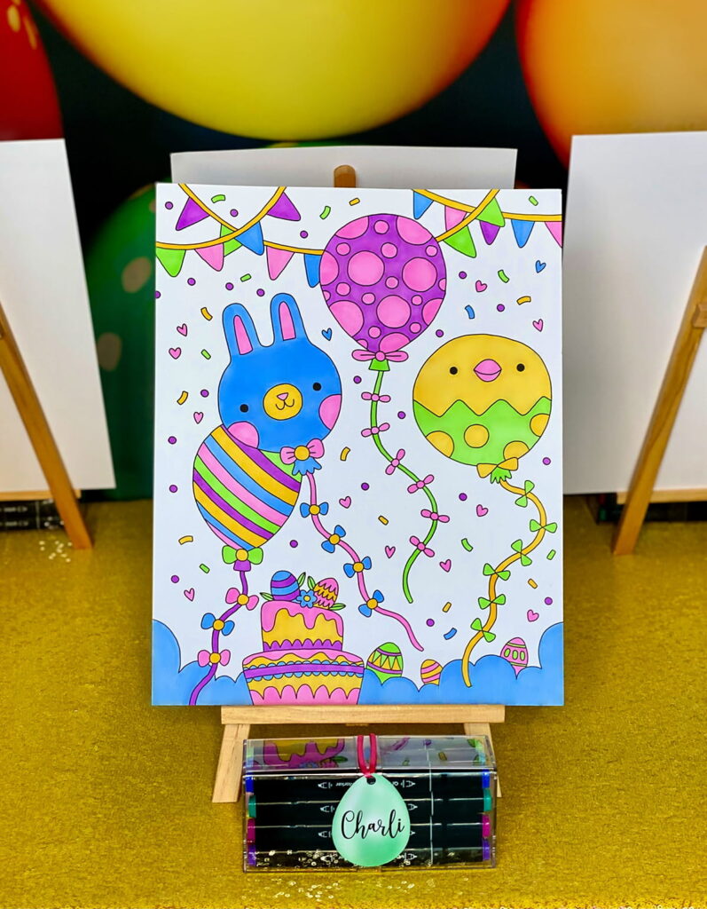 Completed coloured Easter poster displayed on an easel with graphic markers in front and an egg-shaped name tag labeled “Charli.”