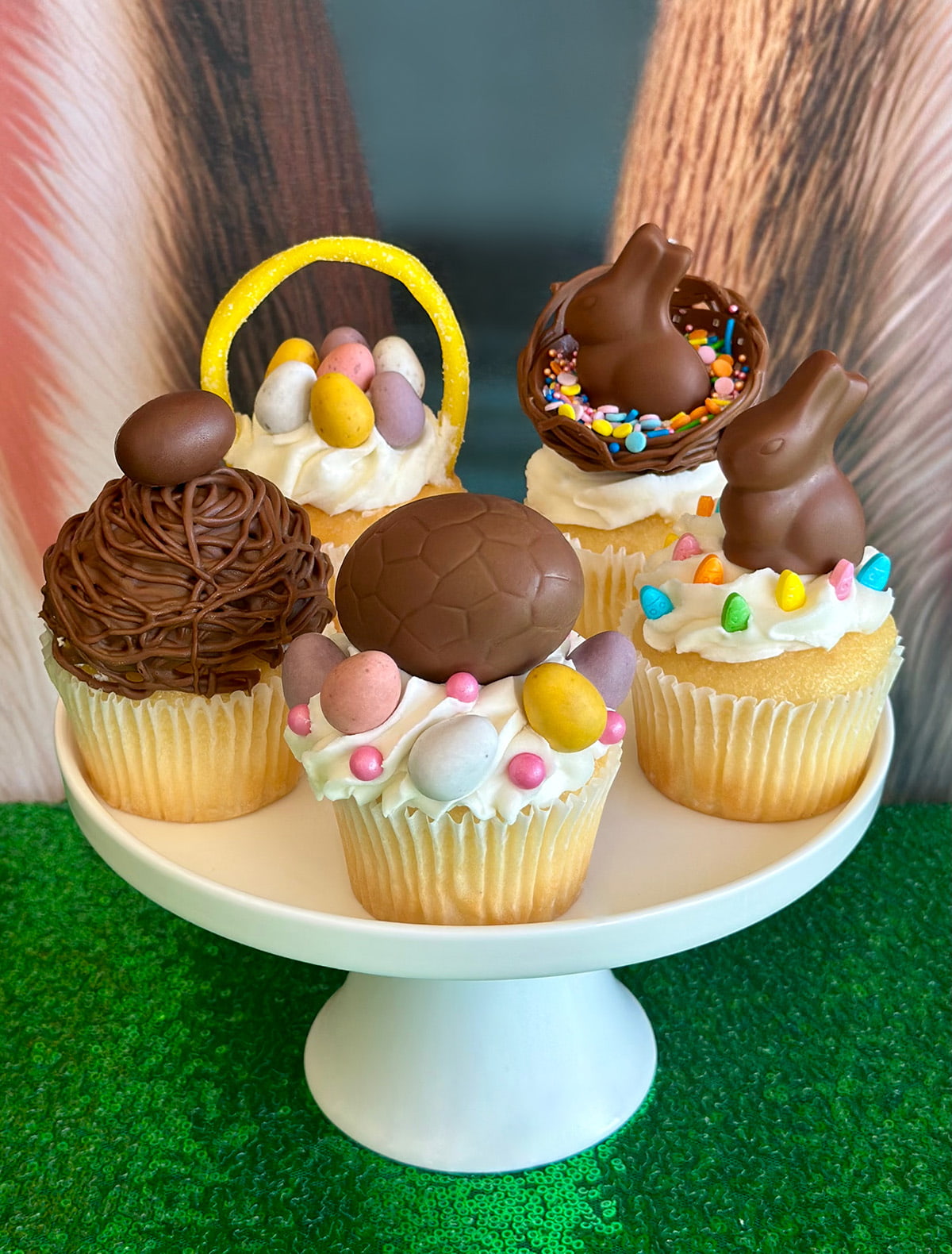 Close-up of one white cake stand holding five decorated Easter cupcakes, including a candy basket cupcake, chocolate bunnies, nests, and sprinkles.