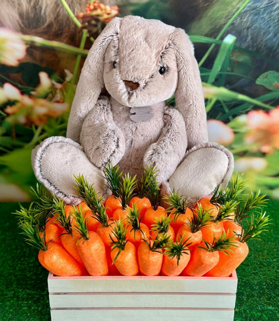 Wooden crate with faux carrots and a plush bunny on top, on a green sequin table with a spring backdrop for an Easter Bunny Adoption party.