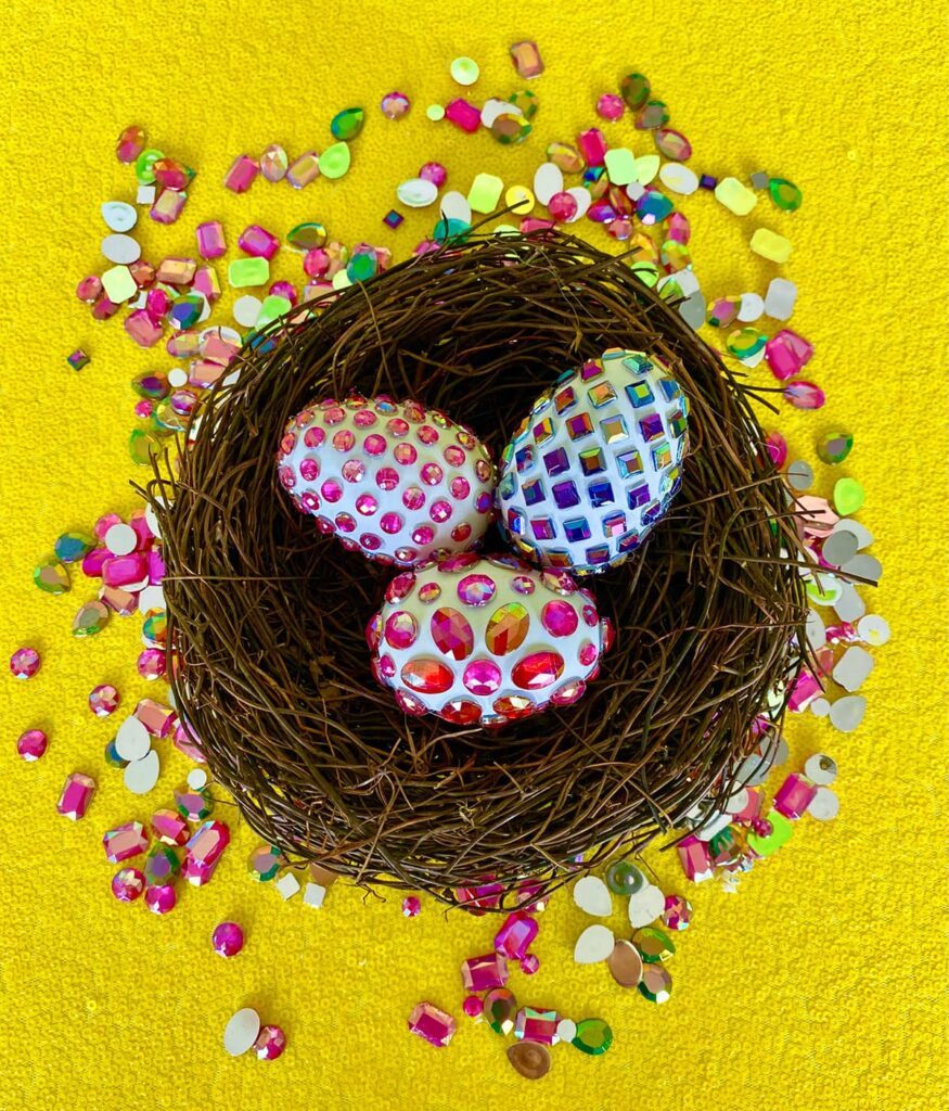 Three gem-decorated eggs resting in a faux brown nest on a yellow sequin tablecloth, with colorful gems scattered around the table.