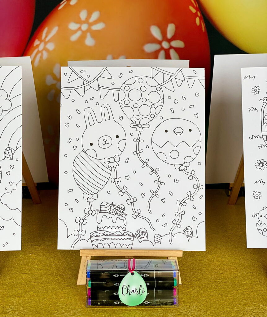 Close-up of an Easter colouring poster on a wooden easel with graphic markers in front, including an egg-shaped name tag labeled “Charli.” Yellow sequin tablecloth and colorful egg backdrop are visible in the background.