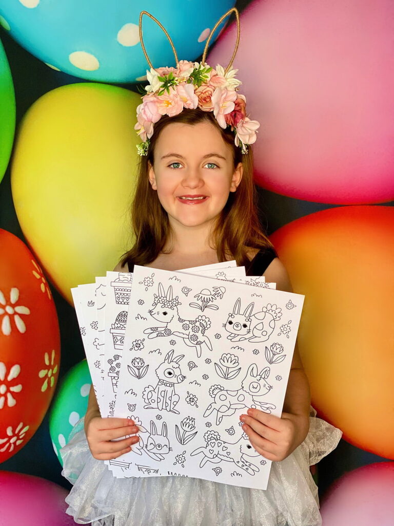 Young girl wearing a floral bunny ear headband, smiling and holding uncoloured Easter-themed posters in front of a vibrant egg backdrop at an Easter colouring party.