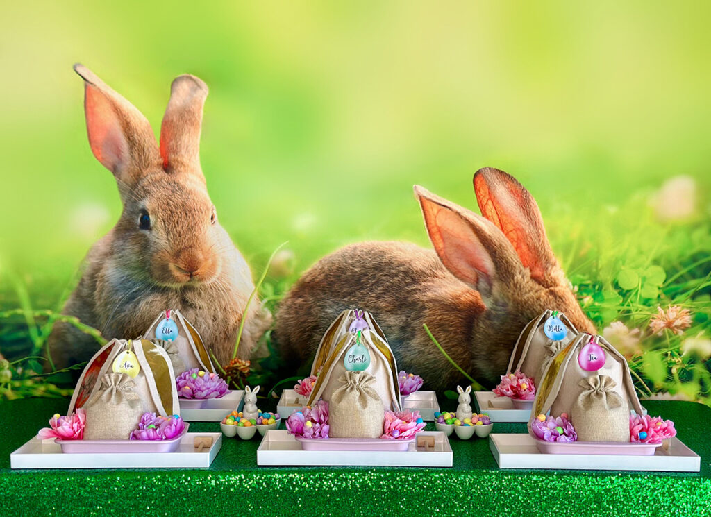 Angled view of a DIY Easter Bunny Bark party table showing five decorated trays with burlap gift bags, silicone pan, pastel flowers, and bunny-egg holders. The setup sits on a sparkly green tablecloth, with the spring bunny backdrop visible behind.