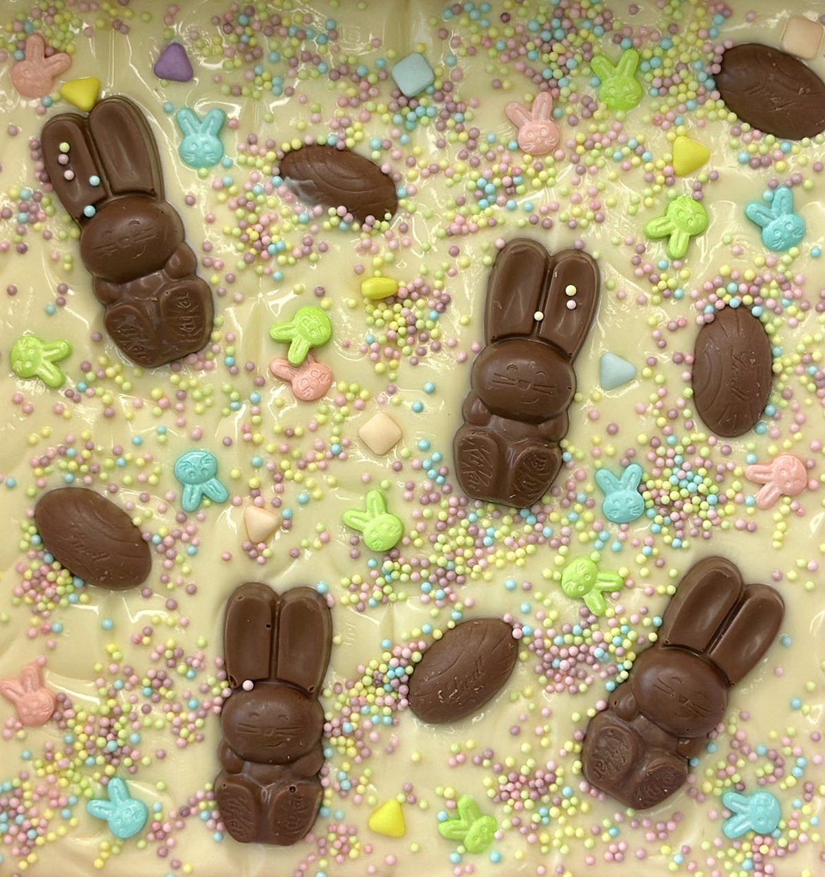Close-up of melted white chocolate topped with pastel Easter-themed sprinkles, mini chocolate eggs, and KitKat bunnies for a kids’ Bunny Bark activity.