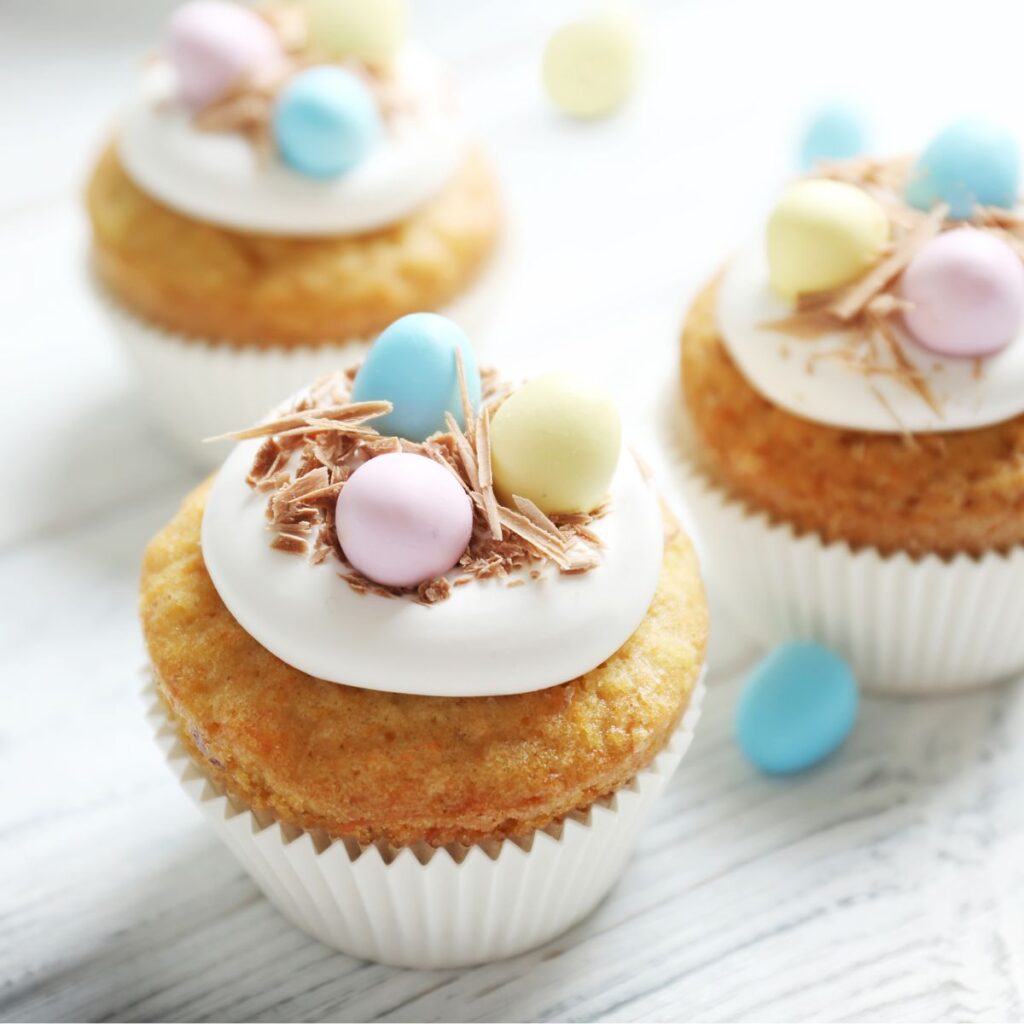 Three cupcakes on a wooden board decorated with white icing, topped with chocolate shards arranged like nests and mini Easter eggs.