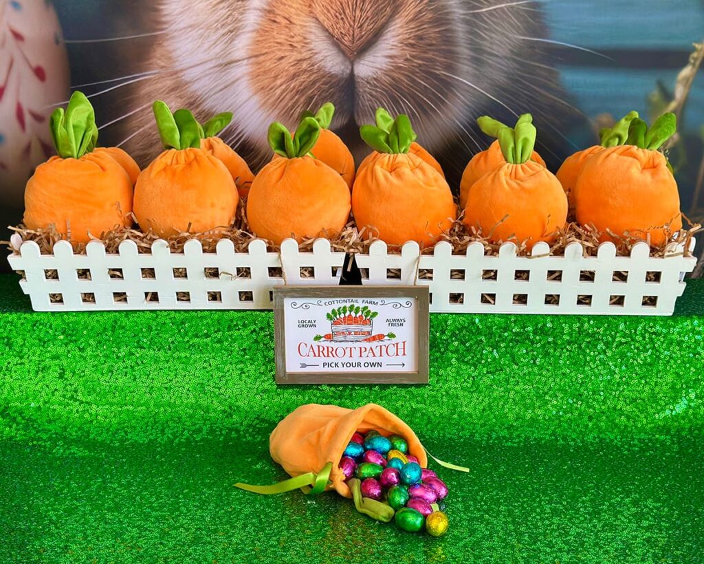 Velvet carrot-shaped treat bags nestled in brown shredded paper inside two white picket garden crates with “Carrot Patch” sign in front; one carrot bag spilling colorful Easter eggs onto green sequin tablecloth, with a bunny-themed party backdrop partially visible behind.