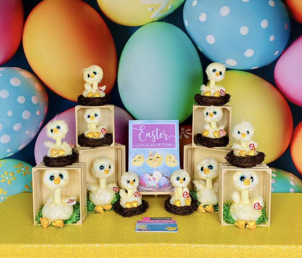 Easter Chick Adoption party setup featuring plush yellow chicks displayed in wooden crates and mini nests on a bright yellow table. A colourful egg backdrop and a central sign reading “Easter Chick Adoption” complete the cheerful spring display.