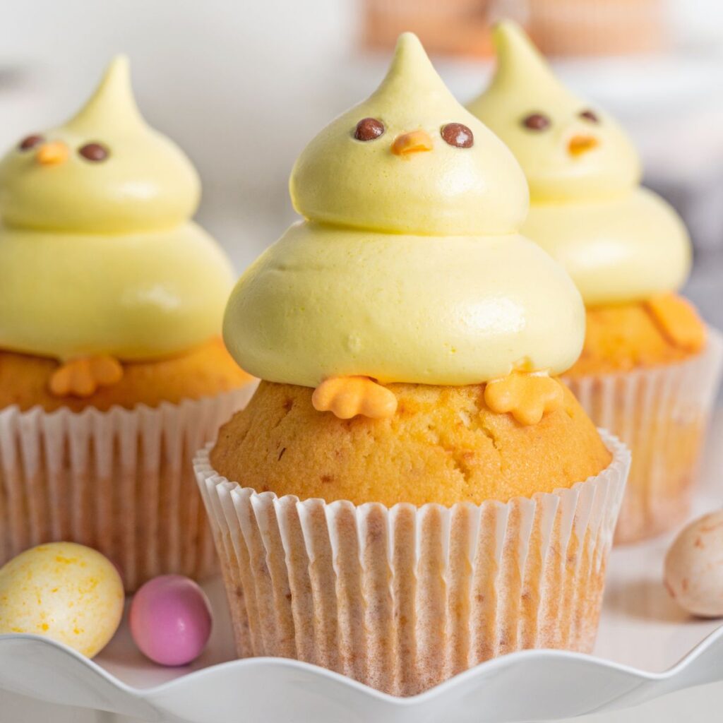 Three carrot cupcakes topped with yellow fluffy icing shaped like plump baby chicks, complete with eyes, beaks, and feet details.