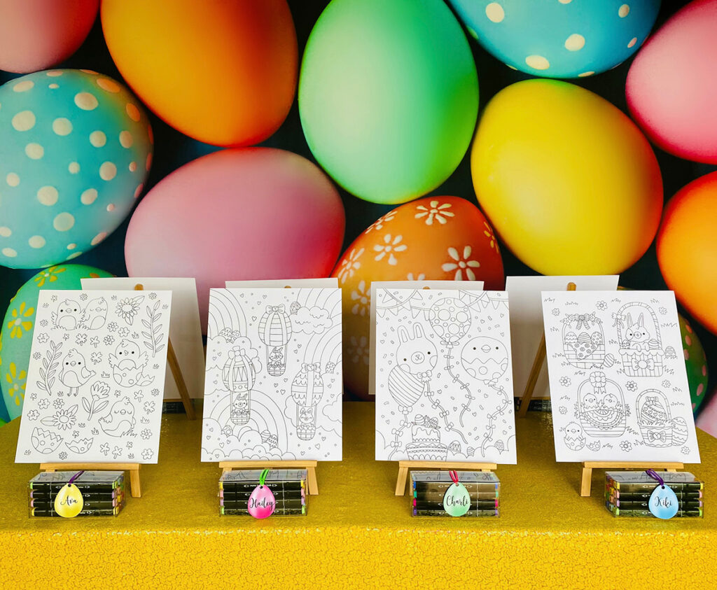 Easter colouring party activity table with a vibrant egg-themed backdrop, yellow sequin tablecloth, wooden easels holding uncoloured Easter posters, and clear boxes of graphic markers with egg-shaped name tags in front.