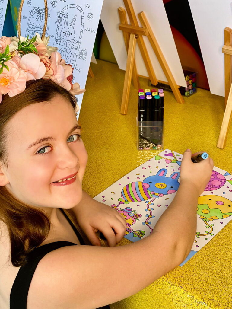 Top-down angled view of young girl looking up at camera while colouring at Easter party activity table with vibrant egg backdrop and yellow sequin tablecloth partially visible.