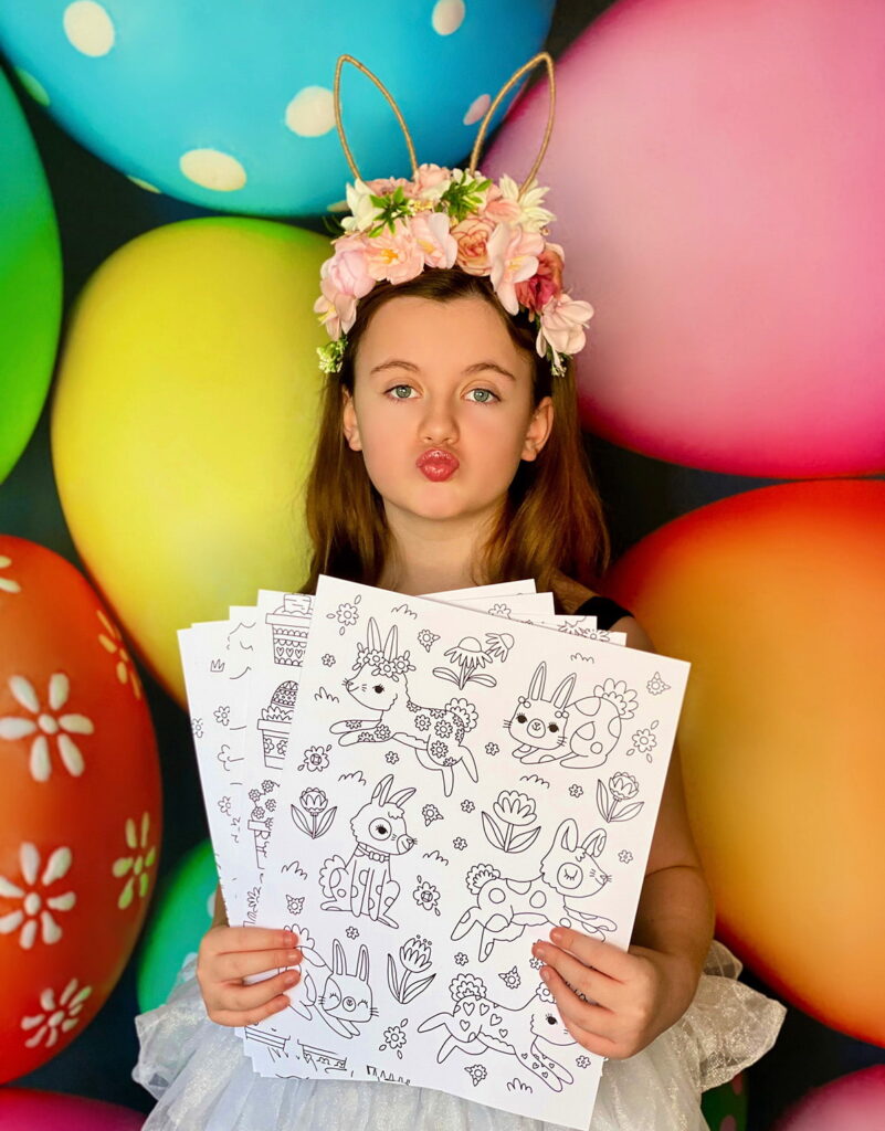 Young girl in front of vibrant Easter egg backdrop making a playful kissy face while holding uncoloured Easter colouring posters.
