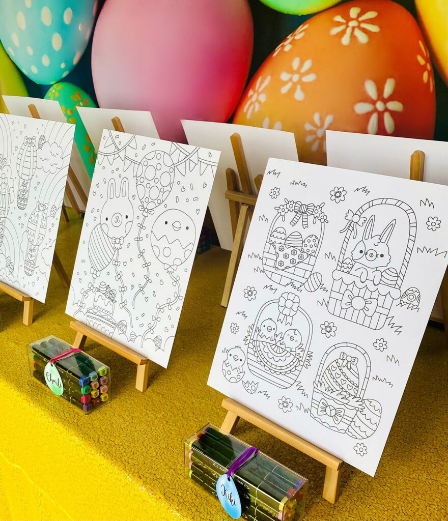 Side view of Easter colouring party table featuring wooden easels with uncoloured Easter posters, clear boxes of markers with egg-shaped name tags, yellow sequin tablecloth, and colorful egg backdrop in the background.