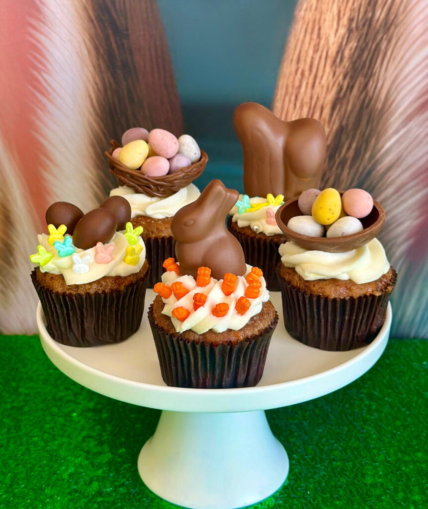 Easter-themed carrot cake cupcakes with cream cheese frosting on a white cake stand, topped with festive decorations like bunny ears, mini eggs, sprinkles, nests, and bunnies, set against a bunny ear backdrop.