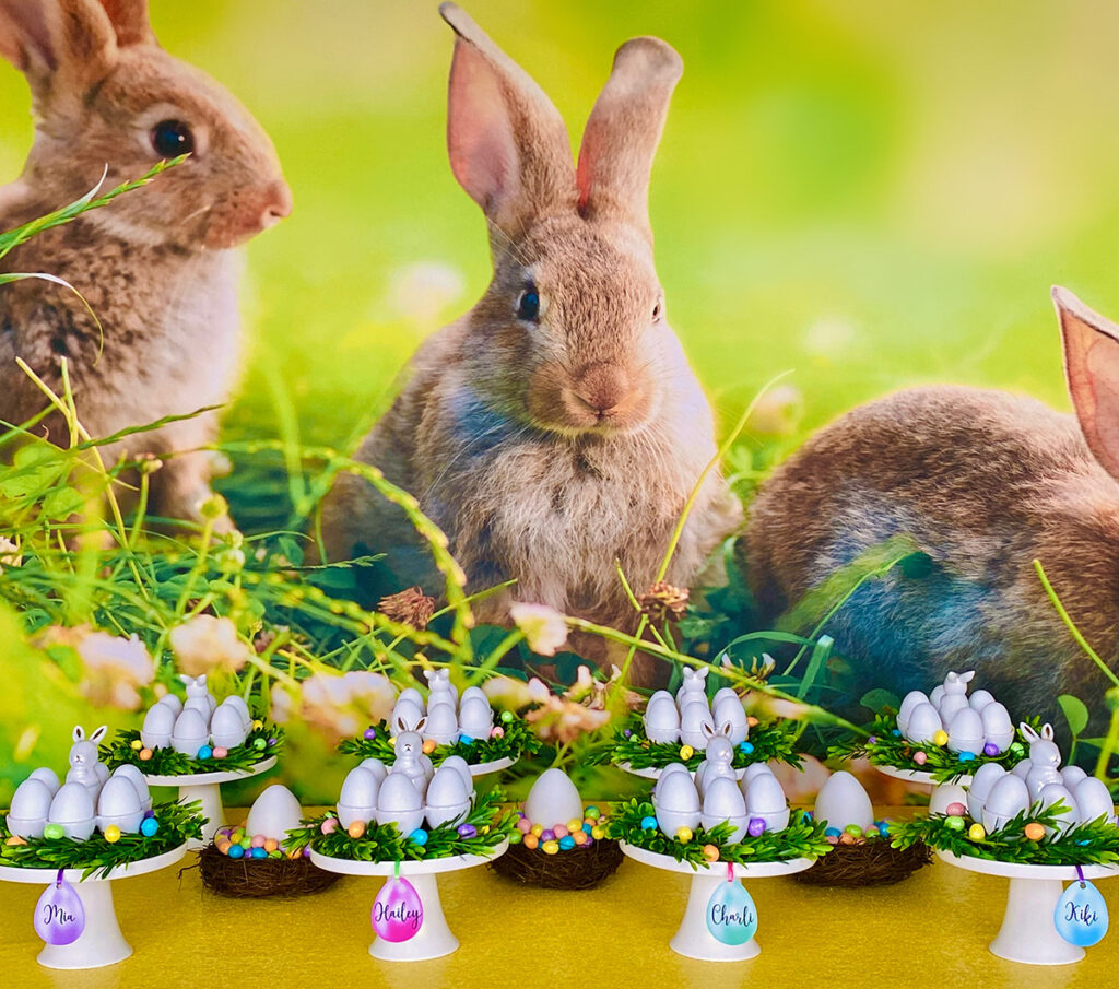 Easter egg decorating party table with a spring bunny backdrop and yellow sequin tablecloth. White cake stands hold bunny egg holders with plastic eggs, surrounded by faux greenery and pastel mini foam eggs. Small nests filled with colourful foam eggs decorate the table.