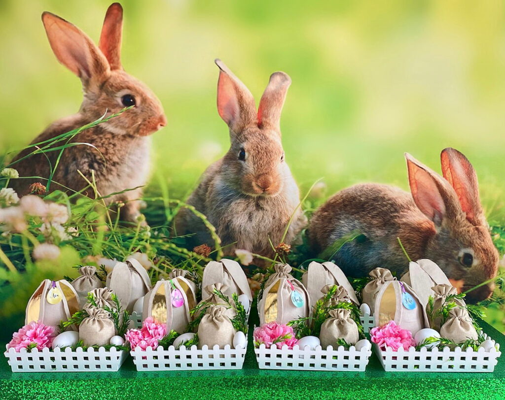 Wide view of an Easter Fairy Garden Party activity table, featuring decorated white crates filled with faux greenery, pastel eggs, and flowers. A bunny backdrop and green sequin tablecloth complete the whimsical spring-themed setup.